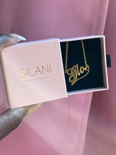 Load image into Gallery viewer, Gold custom name necklace with the name “Glo” and heart detail, placed inside a pink Silani jewelry box, held in hand against a blush pink background.