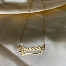 Load image into Gallery viewer, Gold custom name necklace with the name “Djenaba” and heart accent, displayed on smooth champagne satin fabric.